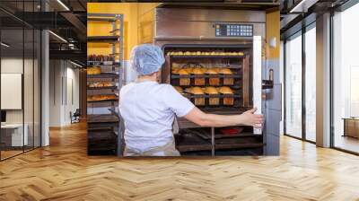 A female baker in white uniform and a beige apron bakes bread and takes cheese buns from an industrial oven. Work at the bakery Wall mural