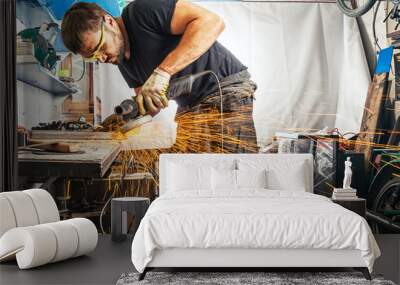 A strong man with hard-working hands in work clothes brewing a metal welding machine on a wooden table in the garage, many sparks fly apart Wall mural