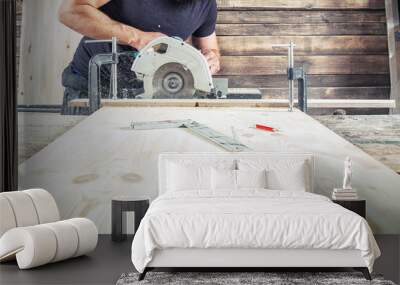 A young brunette man in a black T-shirt is sawing a tree with a modern circular saw in the workshop Wall mural