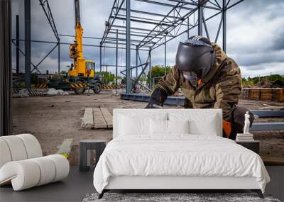 A young  man welder in brown uniform, welding mask and welders leathers, weld  metal  with a arc welding machine at the construction site, blue sparks fly to the sides Wall mural