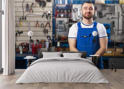 Handsome young man auto mechanic in special uniform clothes is standing and smiling on the background of racks and a desktop with tools for car repair.  Wall mural