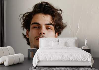 A young man with long curly brown hair and light stubble, looking at the camera with a neutral expression, wearing a white t-shirt, light textured background Wall mural