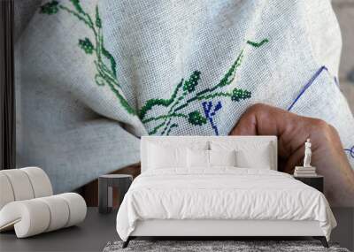 Hands of an elderly woman embroidering a cross-stitch floral pattern on linen fabric. Embroidery, handwork, needlecraft concept. Closeup Wall mural