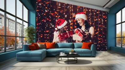 Portrait of cheerful beautiful mother and daughter in Santa Claus hats unwrapping Christmas or New Year s presents against bright garland. Wall mural