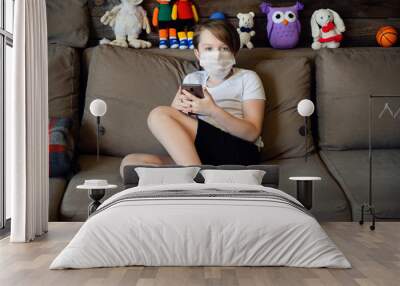 Photo in home interior of a 9 years old boy on a sofa in a medical mask with a phone tablet in his hands in quarantine Wall mural