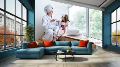 Mom and daughter are sitting by the window and drinking tea, dressed in a white robe and a white towel on their heads. Wall mural