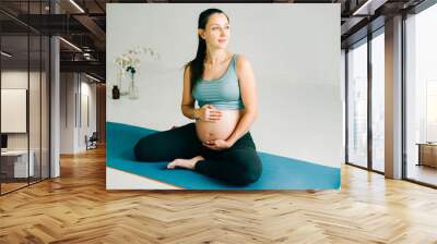 beautiful pregnant girl in sportswear sitting on a yoga Mat and stroking her belly on a studio Wall mural