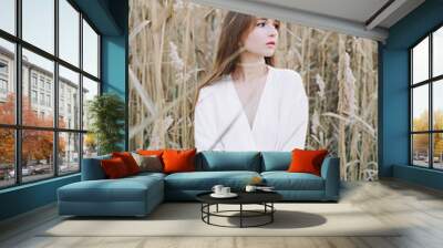 portrait of a young beautiful girl in a white dress, a warm knitted cardigan and black shoes among dry fluffy reeds in the autumn time at sunset Wall mural