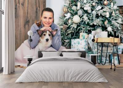 Happy woman with her dog greets, merry Christmas and New Year. The female sits with her pet by the Christmas tree Wall mural