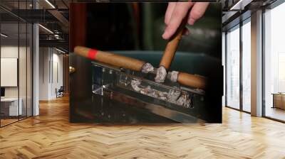 two smoking cigars lie in a transparent ashtray, a man brings a third cigar to an ashtray to knock down the ashes Wall mural