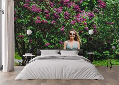 A beautiful curly woman in a summer dress runs and rejoices, she is wearing sunglasses. Against the background of a purple lilac bush Wall mural