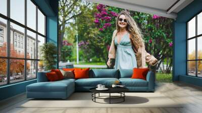 A beautiful curly woman in a summer dress runs and rejoices, she is wearing sunglasses. Against the background of a purple lilac bush Wall mural