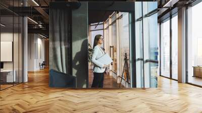 A business woman with long hair in a white jacket and glasses stands with a laptop in her hands in the office space near the window and looks at the city. Work in a modern office with large windows. Wall mural