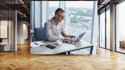 A businessman works on a laptop, a manager sits at a table in the office, works on a laptop. Freelancer workplace, employee at remote work. Remote workplace in coworking, cafe Wall mural