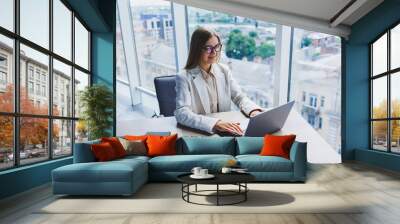 A female head of a company in glasses sits at a laptop in her office with a breathtaking view of the city. Business lady works at a wooden table. Wall mural