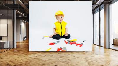 A little boy 2 years old in a builder's suit on a white background. A child in a plastic helmet and plastic tools. Plastic children's toys. Wall mural