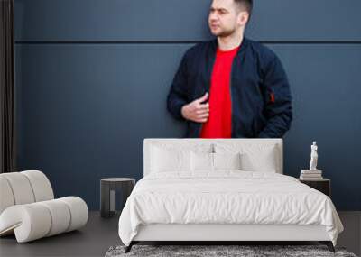 A young man dressed in casual style, a black jacket, jeans, a red T-shirt. He is against a gray wall Wall mural