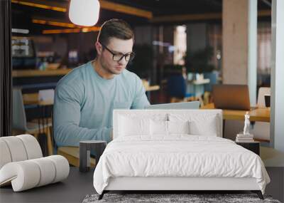 A young promising man in casual clothes sits at a table and works on a laptop. Work space for remote work. Wall mural