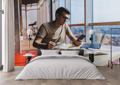 Handsome man in red casual t-shirt writing a message on mobile phone while sitting near glass window at table with laptop working remotely in cafe Wall mural