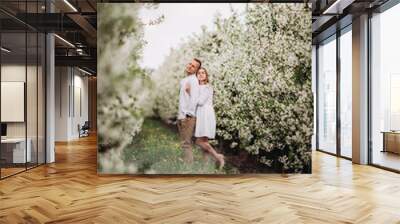 Happy family couple in spring blooming apple orchard. Young couple in love enjoy each other while walking in the garden. The man holds the woman's hand. Family relationships Wall mural