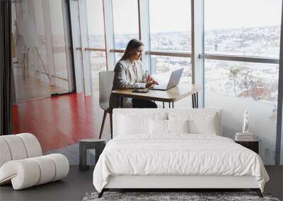 Image of a happy young woman in a jacket smiling and working on a laptop while talking on the phone in a modern office with large windows. Remote work Wall mural