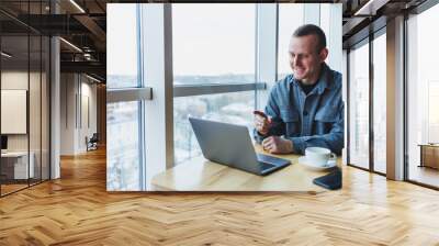 Successful happy business man is sitting at a table in a cafe, holding a cup of coffee and using a laptop. Wall mural