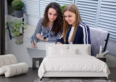 Two successful young business women manager are sitting at a table with a laptop and a notebook working on a new development project, female students write a report on the work of their computer Wall mural