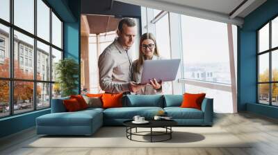 Two young businessmen and businesswomen smiling while sitting in the office, talking and surfing the internet together with a laptop Wall mural