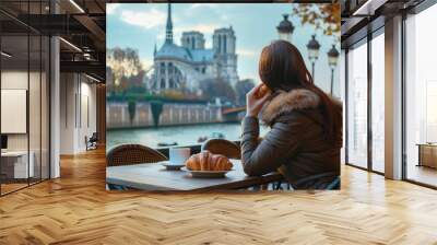 A girl sitting at a cafÃ© table, with a croissant in hand and the Notre-Dame Cathedral in the background Wall mural