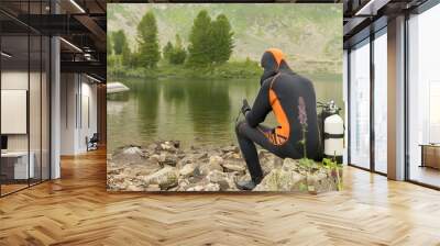 a guy in a diver's suit for diving in the water, next to oxygen tanks, sits on rocks on the lake shore against the background of a mountain-forest landscape against the background of a passing boat Wall mural