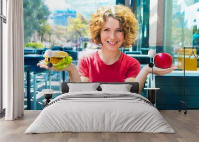 Beautiful girl, young happy woman holding a ripe red apple and burger on the palms, choosing between, smiling. Junk unhealthy fast food in one hand versus vs healthy in other. Difficult right choice. Wall mural