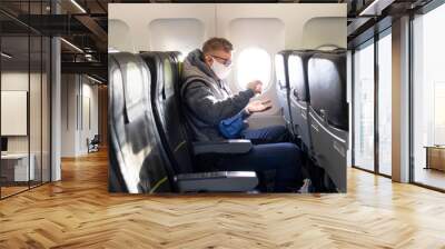 Guy in airplane, young man in glasses, medical protective sterile mask on his face sitting on plane using, apply anitizer for disinfect hands against coronavirus, virus bacteria. Pandemic covid-19 Wall mural