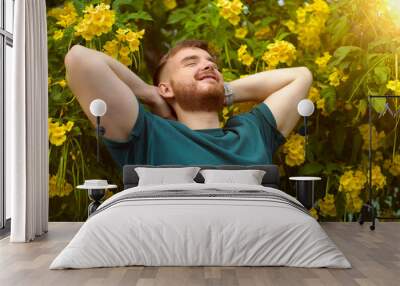 Portrait of happy handsome bearded guy, young positive man with beard is smelling beautiful yellow flowers in the garden, smiling, enjoying spring or summer day, breathing deep deeply fresh air Wall mural