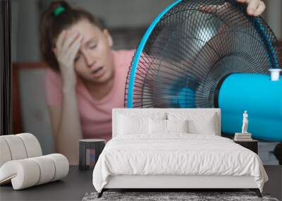 Young woman using electric fan at home in living room, sitting on couch cooling off during hot weather, suffering from heat, high temperature Wall mural