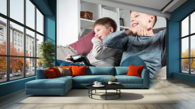 Portrait of two laughing teenage boys children sitting on grey sofa at home, holding gaming controller joystick, playing videogames, raising hands, gesturing. Hobby, free time, gaming, friendship. Wall mural