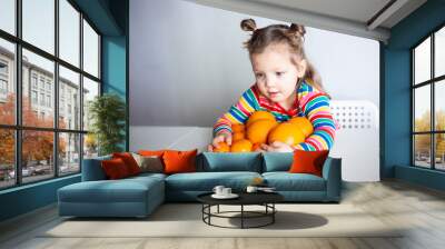 Little girl, 3 year old baby, with a ponytail hairstyle in a colorful colorful striped jacket hugs a lot of appelsins Wall mural