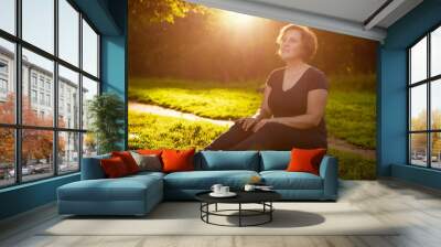 Adult woman with short hair sitting in a dark dress in the park on the grass in the rays of the setting sun summer day Wall mural