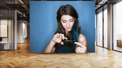 Beautiful brunette woman with a smile cutting long hair with scissors on a blue background. Happy girl Wall mural