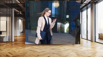 Business woman of Caucasian ethnicity in a coat and a suit with glasses writes SMS messages on the phone near the office building. Business people distance online communication concept Wall mural