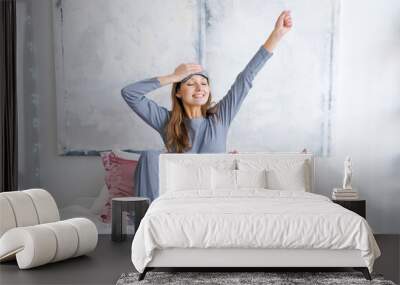 Happy young woman sitting in comfortable bed, arms outstretched after waking up in the bedroom, wearing blue pajamas with a sleep mask, doing exercises in the morning, welcoming a new day, sunrise Wall mural