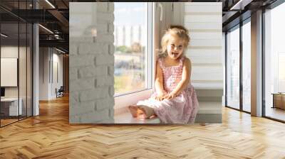 beautiful little girl in a pink dress sits at the window on a sunny day and smiles Wall mural