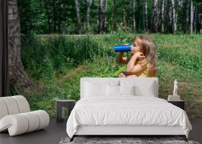 little girl drinking soda from a metal can by the lake while sitting on the grass on a hot summer day Wall mural