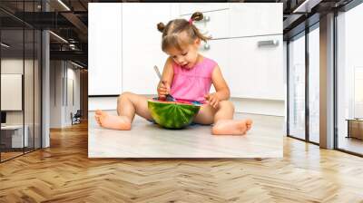 little girl eating watermelon with a spoon while sitting on the floor at home Wall mural