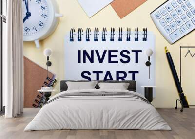 On a light background, a white alarm clock, a calculator, a pen and a notebook with the text INVEST START. Flat lay Wall mural