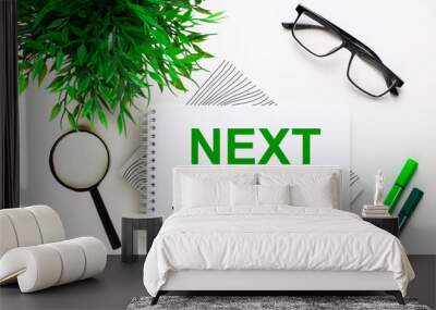 On a white background lies a notebook with the word NEXT STEPS, glasses, a magnifying glass, green markers and a green plant Wall mural