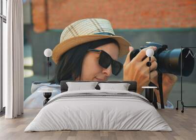 Young Caucasian woman with photography camera, wearing white shirt, hat and sunglasses takes pictures on city street. Wall mural