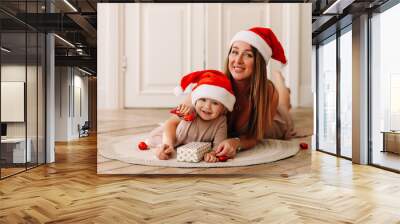 Cute smiling mom and little baby daughter in Santa hats hugging having fun laughing lying on the wooden floor waiting for the Christmas holiday and New Year, selective focus Wall mural