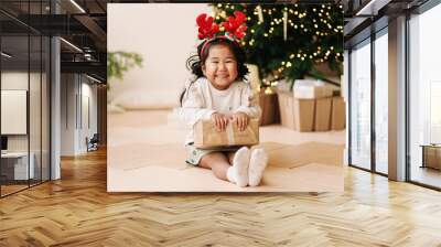 Portrait of a cheerful happy positive emotional Asian little girl the child is having fun rejoicing and holding a gift box on a Christmas holiday in a decorated house during the New Year vacation.  Wall mural