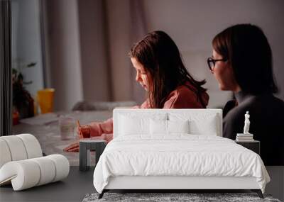 A focused young girl paints carefully on paper at a table, with an adult female figure observing in the background Wall mural