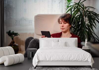 A tranquil scene unfolds as a woman engrossed in her e-reader rests in a comfy armchair, basking in the gentle light of her cozy nook Wall mural